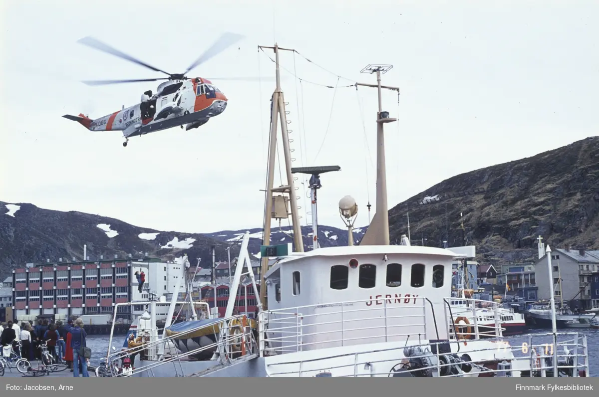 Et Sea-King redningshelikopter demonstrerer redning i Hammerfest indre havn. To personer henger i wiren under helikopteret. En liten gruppe mennesker står på kaia og ser på og noen tråsykler og en moped bak dem. FFR-båten Jernøy ligger ved kai med landgangen ute. Bak hekken til Jernøy ses deler av Findus-anlegget og Mollafjellet i bakgrunnen. Foran rorhuset til Jernøy ligger FFR-båten Brynilen ved Bangkaia. Til høyre for den ligger en stor fiskebåt med en liten plastbåt inntil. I bakgrunnen er fjellet Salen. Noen snøflekker ligger enda på Mollafjellet en tidlig sommerdag.