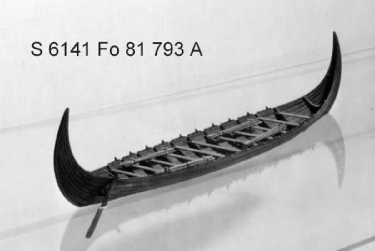 Fartygsmodell av oxelträ, rekonstruktion av "Stora Kvalsundsbåten, utgrävd 1920", daterad till 400-700 e.Kr.. Skrov utfört i två halvor med limfog genom köl och stävar. Bordläggningen skuren på såväl in- som utsidan. Klamparna på borden pålimmade. Håarna av lövträkvistar, med självvuxen knagge, fastbundna med svartoxiderad suturtråd. Skrovet betsat i tjärfärg.