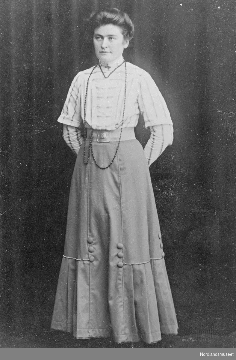Fagfoto av 1 kvinne i helfigur kledd i lys bluse og mørkt skjørt. Rundt halsen henger et langt smykke.