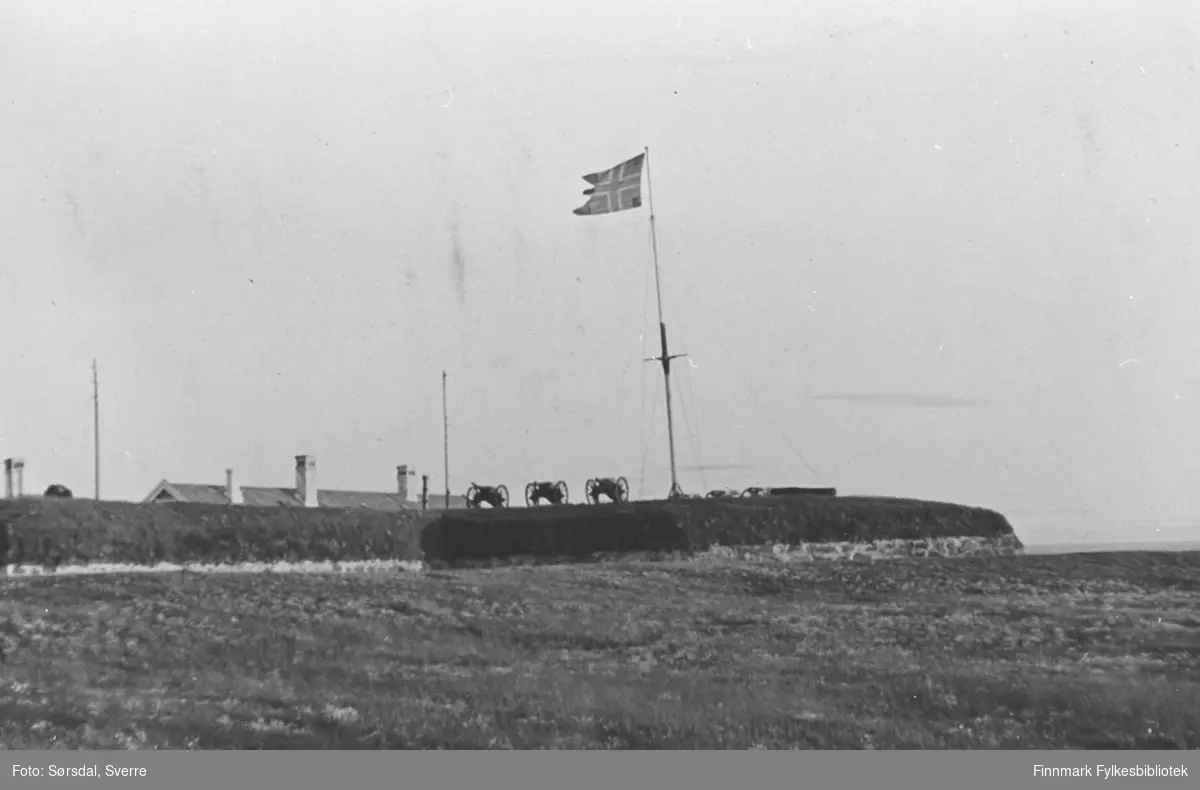 Bildet av Vardøhus festning med flagget til topps. Vi ser festningsvollene og tre putalova saluttkanoner. I bakgrunnen sees taket til kommandantboligen.