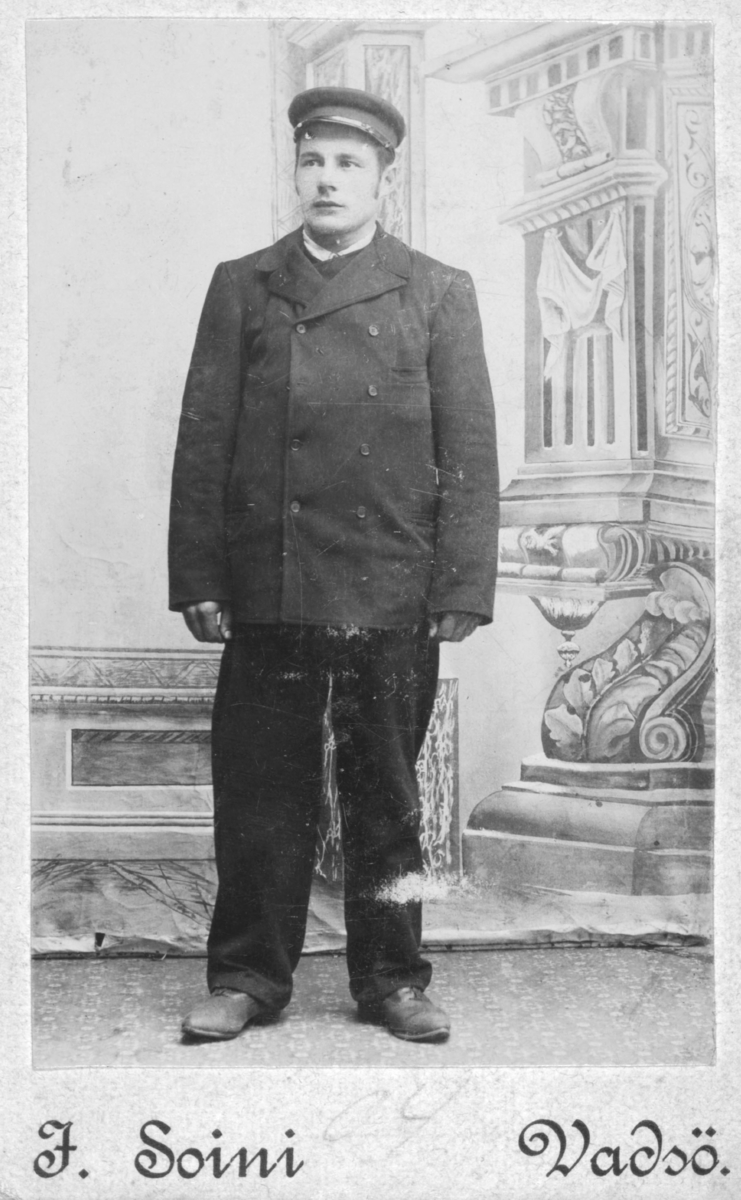 Et portrett av en ung Werner Alfred Korbi. Han er kledd i jakke, skjorte, bukser, hansker, sko og hatt. I bakgrunnen kan man se en malt kulisse.