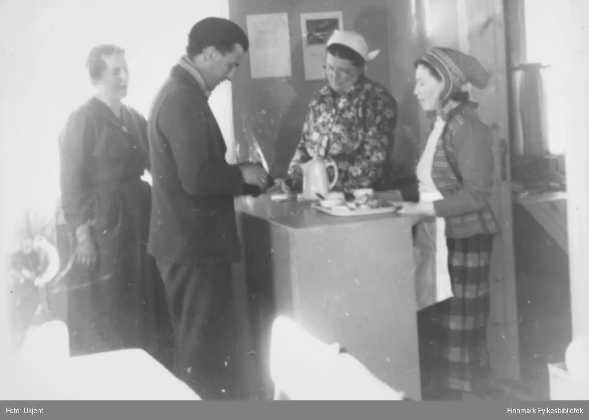 Bildet er fra åpningen av Kautokeino husmorlagets kafé i Kautokeino i 1955. Fra venstre: Anna Jonsen, Oddmund Sandvik, Hilma Gjertsen og Ynga Turi Hoffa. Oddmund Sandvik, mangeårig ledende politiker, var første gjest. Ved siden av kafeen rommet det nye bygget et vaskeri og utleielokale av to klasserom fram til nyskolen ble åpnet i 1957. Luftforsvarets stasjon med vaskingogstell av uniformer m.m og sengetøy til et mannskap på vel 100 personer var ensentral kunde og oppdragsgiver for det nye vaskeriet.

Påskrift på bildet er: "Husmorskurs, kafe åpnet år 53-54. På bilde Anna Jonsen, Oddmund Sandvik, Ynga Turi Hoffa og undertegnedes mor Hilma Gjertsen". Ynga har på seg en samisk lue og et forkle. Ved siden av henne har Hilma på seg et skaut og briller. Kvinnene har på seg skjorter og skjørt. Oddmund har på seg et skjerf, jakke og bukse. 

Bildet er fra åpningen av Kautokeino husmorlagets kafé i Kautokeino i 1955 der Oddmund Sandvik, mangeårig ledende politiker, var første gjest. Ved siden av kafeen rommet det nye bygget et vaskeri og utleielokale av to klasserom fram til nyskolen ble åpnet i 1957. Luftforsvarets stasjon med vaskingogstell av uniformer m.m og sengetøy til et mannskap på vel 100 personer var ensentral kunde og oppdragsgiver for det nye vaskeriet.