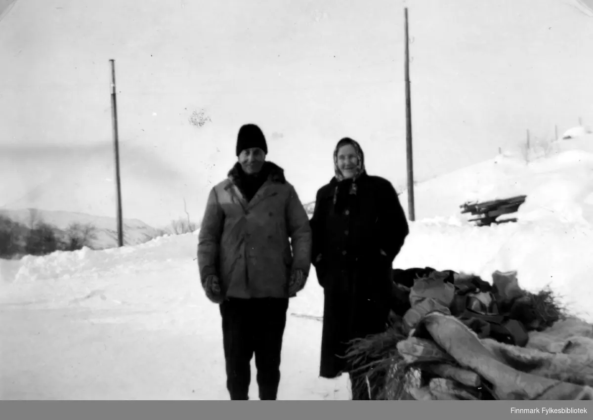 Fotografi av Abiel og Jenny Randa. De står ute, ved siden av en slede. Det er vinter og det ligger mye snø på bakken. Det står to stolper bak dem. Abile er kledt i jakke, bukse og votter. På hodet har han en mørk lue. Jenny er kledt i en mørk kåpe. Hun har et skaut på hodet