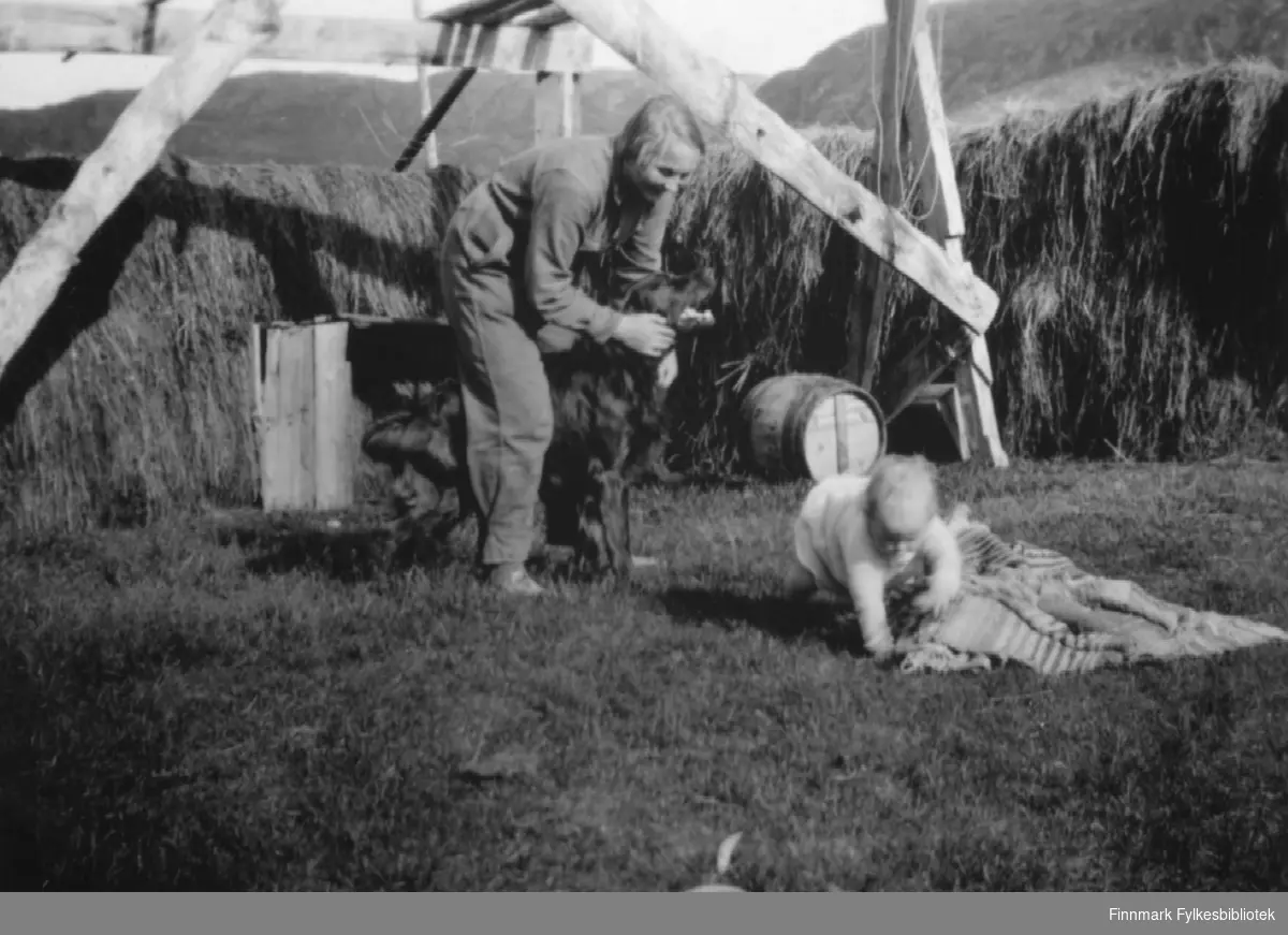 Ella Gunnari holder hunden, mens Pia Gunnari boltrer seg i gresset, sommeren 1935. Kanskje har de seg en liten pause frå slåttonna, i bakgrunnen henger i hvertfall slåttehøyet til tørk.