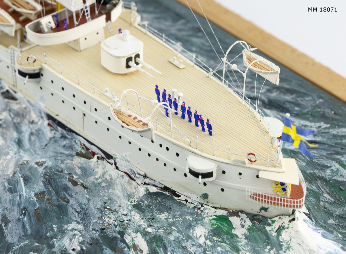 Fartygsdiorama visande två fartyg under gång till sjöss. Modellen är tillverkad av trä med detaljer av plast, papper och metall. Modellerna är placerade i ett hav av någon form av plastelina, målat i gröna och blå toner. Havet är uppbyggt på en rektangulär platta. Modellerna är detaljerat återgivna och visar pansarkryssaren "Fylgia", den ena modellen visar fartyget i sitt ursprungliga skick, den andra modellen visar fartyget efter moderniseringen 1939-40. Den "äldre" modellen visar ett vitt fartyg med rök bestående av grå ull från de tre skorstenarna, den "yngre" modellen visar ett kamouflagemålat fartyg försett med vitt neutralitetsband. Dioramat är signerat på havet med rött: "H Biärsjö".