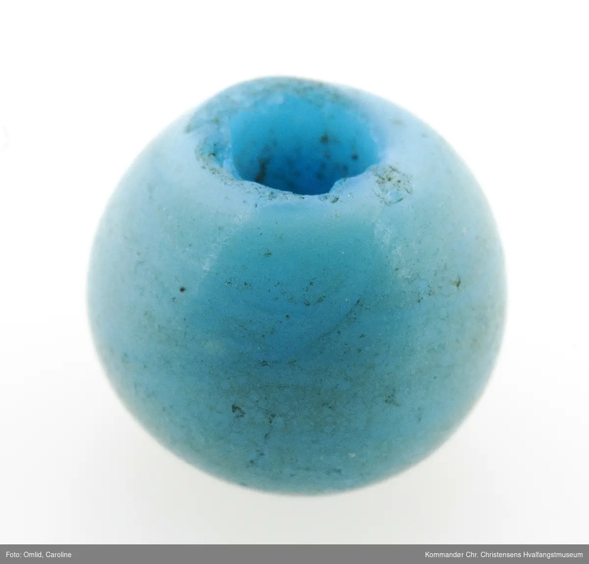 En lys blå glassperle, med innsklag av dypere farge. Hull i perlen.
