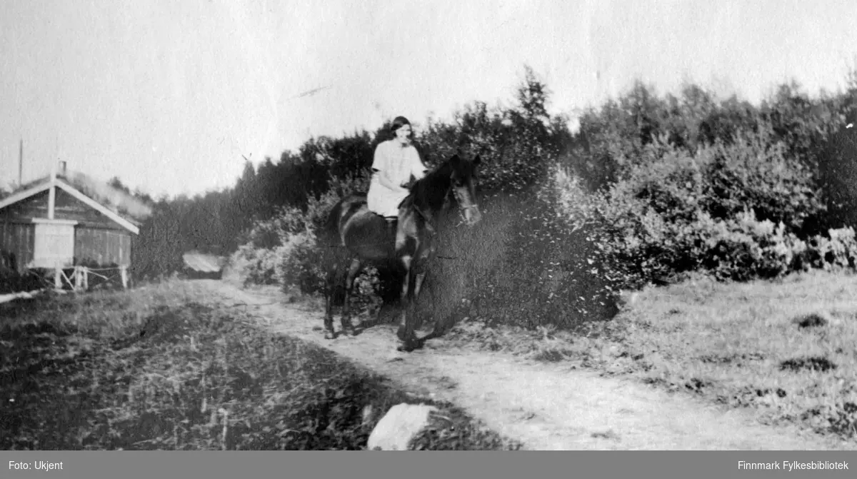 Bildet er tatt fra gården Strømsnes i Jarfjord sommeren 1924. Elsa Bertelsen rir langs gårdsveien på hesten Båro. Elsa har på seg en kjole. Bak henne og hesten kan man se et hus med torvtak. Huset er bygget av tremateriale. På deres høyre side kan man se skog, planter og busker. På venstre side av stien kan man se en stor sten.