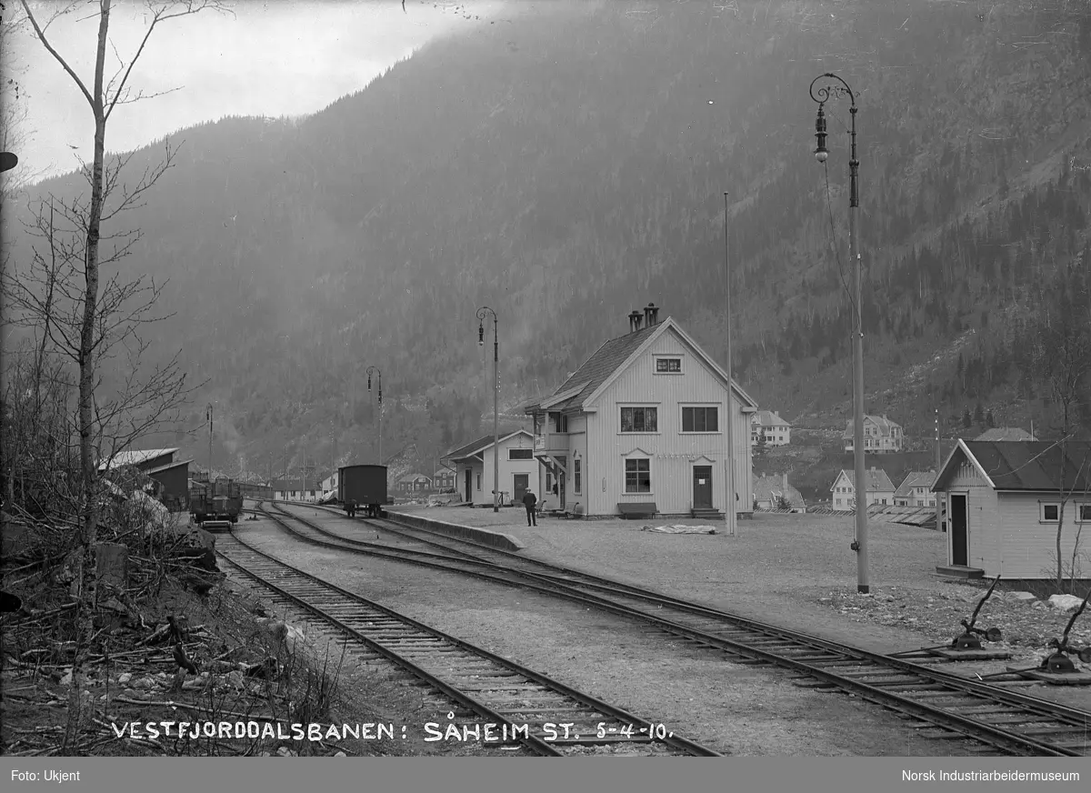 Såheim stasjon (Rjukan stasjon) med lukket godsvogn av typen G3 stående foran pakkhus på stasjonstomten og en kassevogn av typen L4 ved siden av bygning mot sør. To sporvekslere langs jernbanesporet. Belysning på fire stolper langs perrongen. Bolighus i Flekkebyen og Villaveien i bakgrunnen.