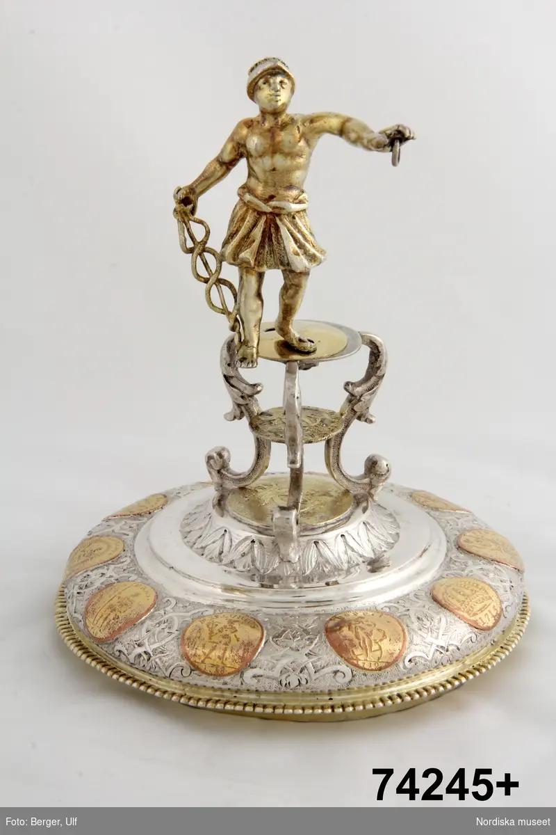 Bägare på låg genombruten gallerfot, nedtill med pärlrand, bägarens sneda del omslutet av ett rikt cislerat ornament  med frukter, blad och band, förgyllda svängda ben, mellan vilka ett karolinmynt från år 1718 , å trefoten en statyett Mesenius, (höjd 8 cm) stående ikl en kort kjortel och hjälm på huvudet, den vänstra armen utsträckt hållande en ring, den högra hållande caducéen stödd mot marken, fana med stång saknas. UPK 1904.
Stämplar: Stockholms stads vapen, åldermansranka, årsbokstaven M andra serien, IB.