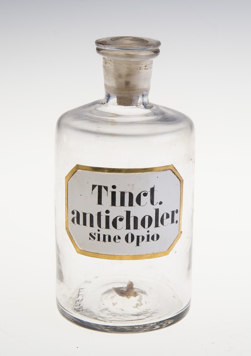 Sylinderformet glassflaske med flat glasspropp, pålimt rektangulær signatur med svart skrift og ramme påmalt i gulllfarge. Flasken har inngravert tallet 26.