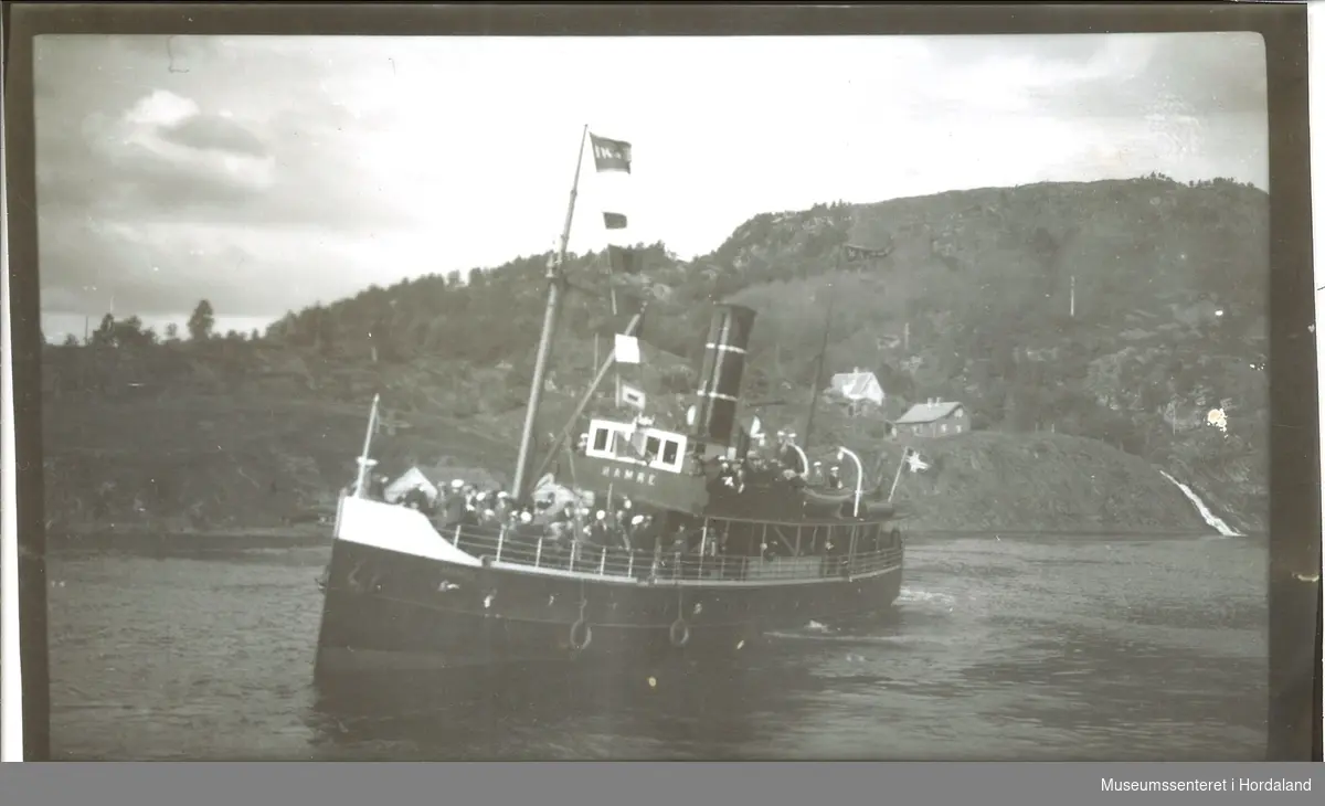 Dampskipet "Hamre" med masse folk. Norskflagg på båten, fjellandskap med små hus i bakgrunnen. Bildet er tatt fra kommunekaien i Fotlandsvåg.