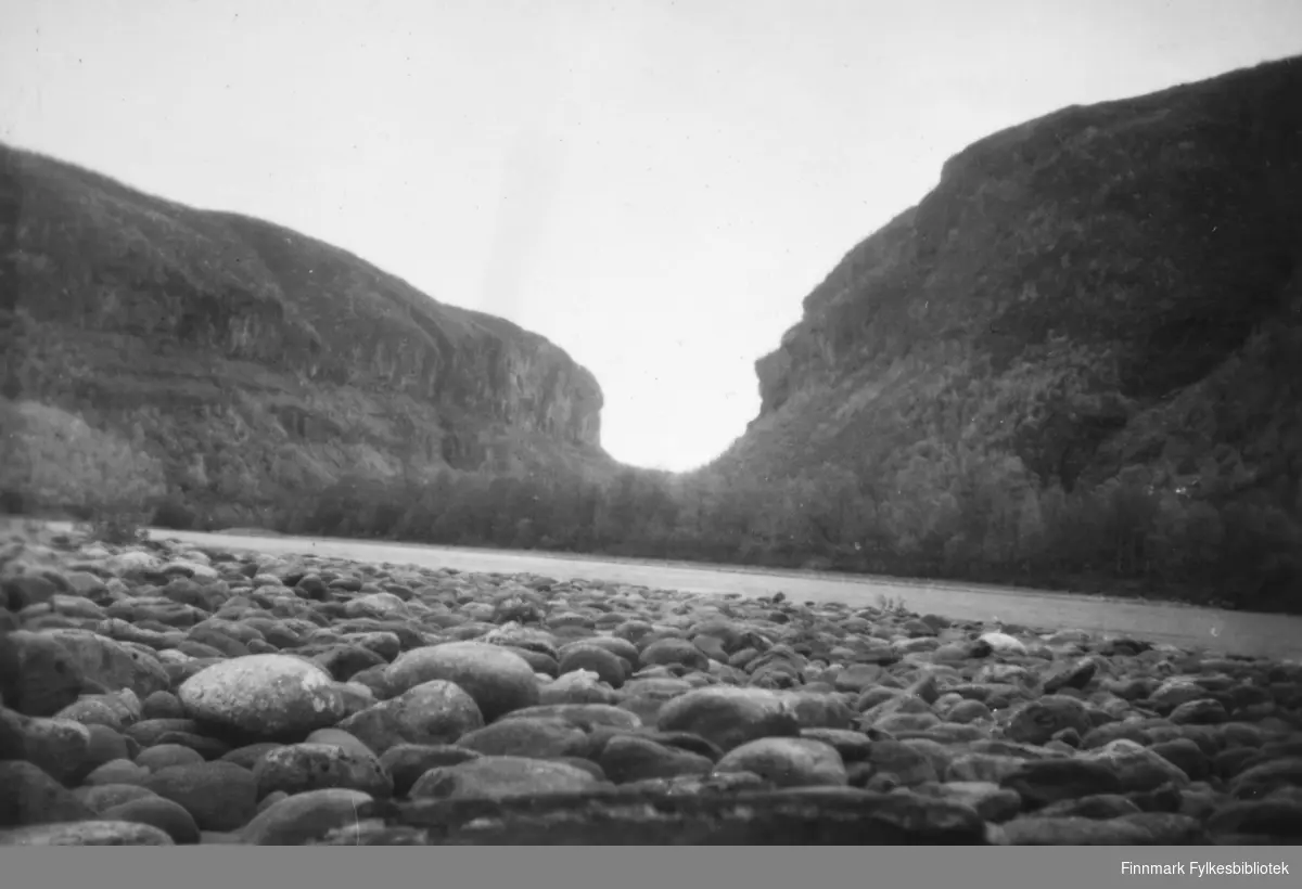 En canyon i Altadalen ses fra Altaelvas bredd. I forgrunnen ser vi en rullesteinstrand. Langs elva ser vi skog. ca.1950-60.