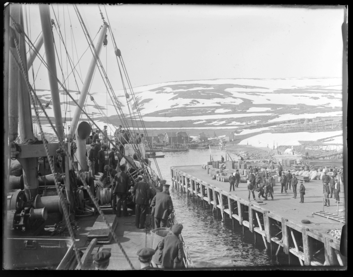 Hurtigruta legger inn til kai ved Båtsfjord ca. 1920. Bildet er tatt ombord i hurtigruteskipet D/S FINMARKEN og viser en del av hurtigruta og kaia, der det står det mange folk og tar i mot anløpet. Sekker, tønner og annet som skal fraktes ligger stablet på kaia.