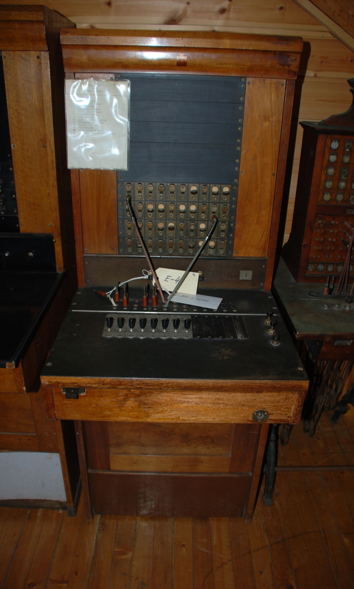 Sentralbord med koblingspanel der telefonsamtaler kobles ved hjelp av kabler med plugg. Står på gulvet og betjenes av en person. Kapasitet 40 linjer, for magnettelefon.