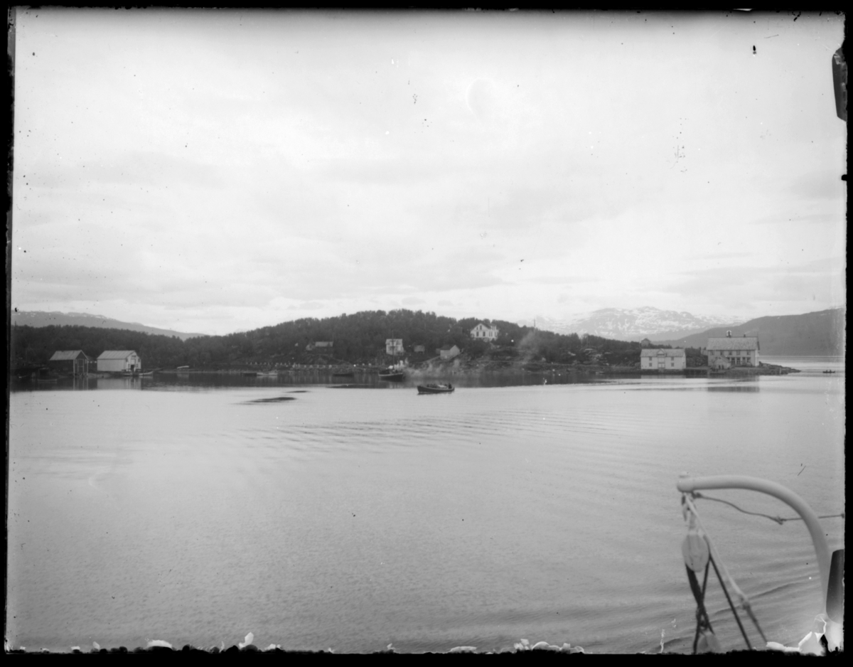 Bildet er tatt fra båt og vi ser en øy (?) med noen hus og sjøboder. Det er trær på øya og snøkledte fjell i bakgrunnen. Bildet kan være fra reisen nordover i 1912. Det ser ikke ut til å være fra Finnmark