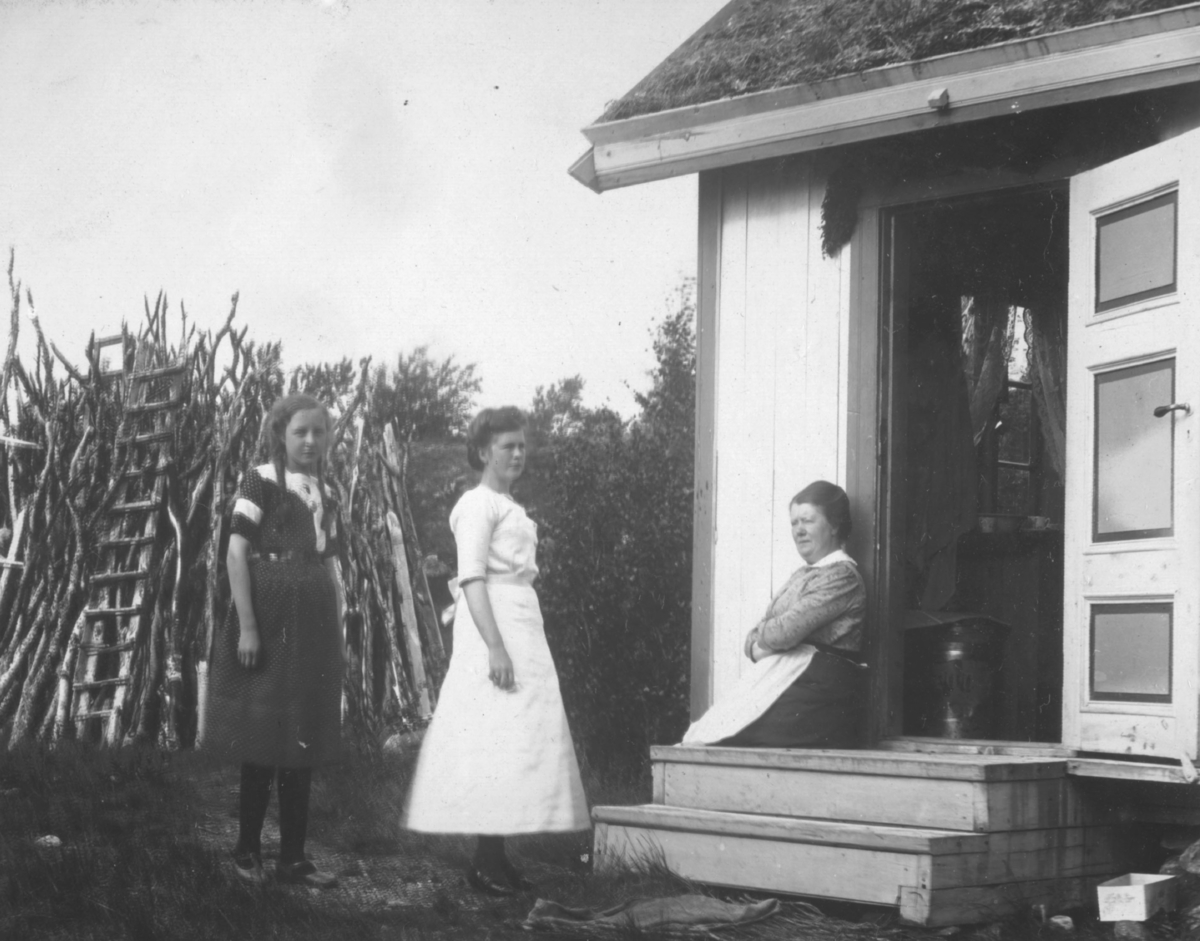 En dame sitter på trappen utenfor hytta. To jenter står ved siden av. Den ene har lys sid kjole, og den yngste fletter og mørk kjole. Bak dem er det et råveskjul.