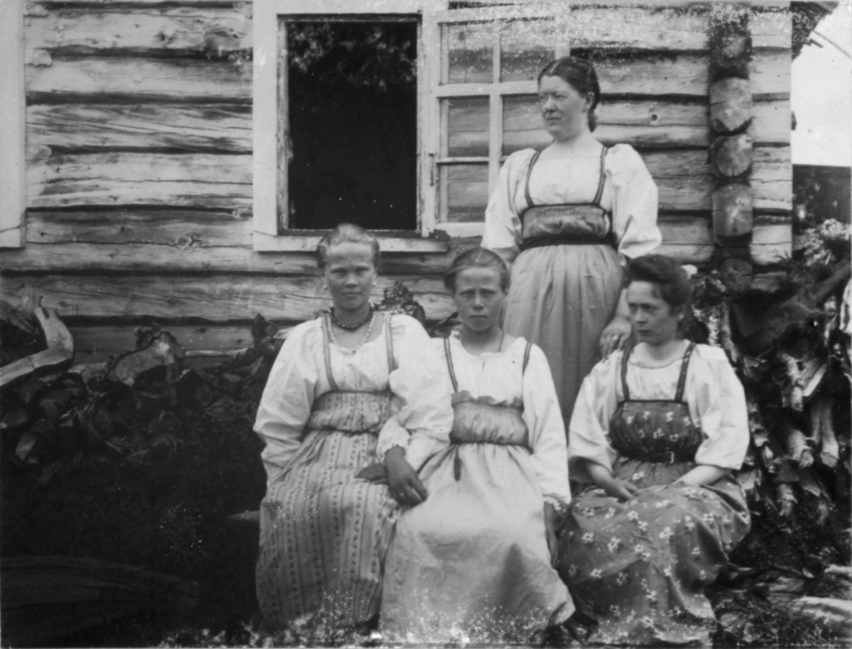 Fire damer foran en bygning. Tre av de sitter, og den ene står bak ved et åpent vindu. Foran huset er det stabler med ved. Damene har lyse bluser og side lyse skjørt med snøring i livet.