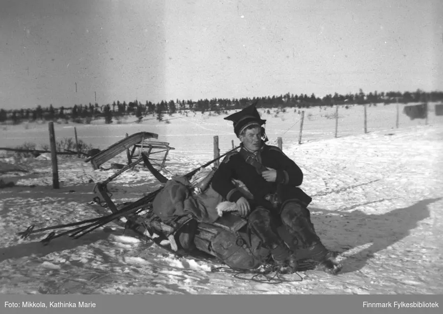 I Pakanajoki, ca. 1946-1947. Andreas Mikkola i samekofte og stjernelue sitter på en fullpakket slede. I bakgrunnen er en pulk stilt på høykant