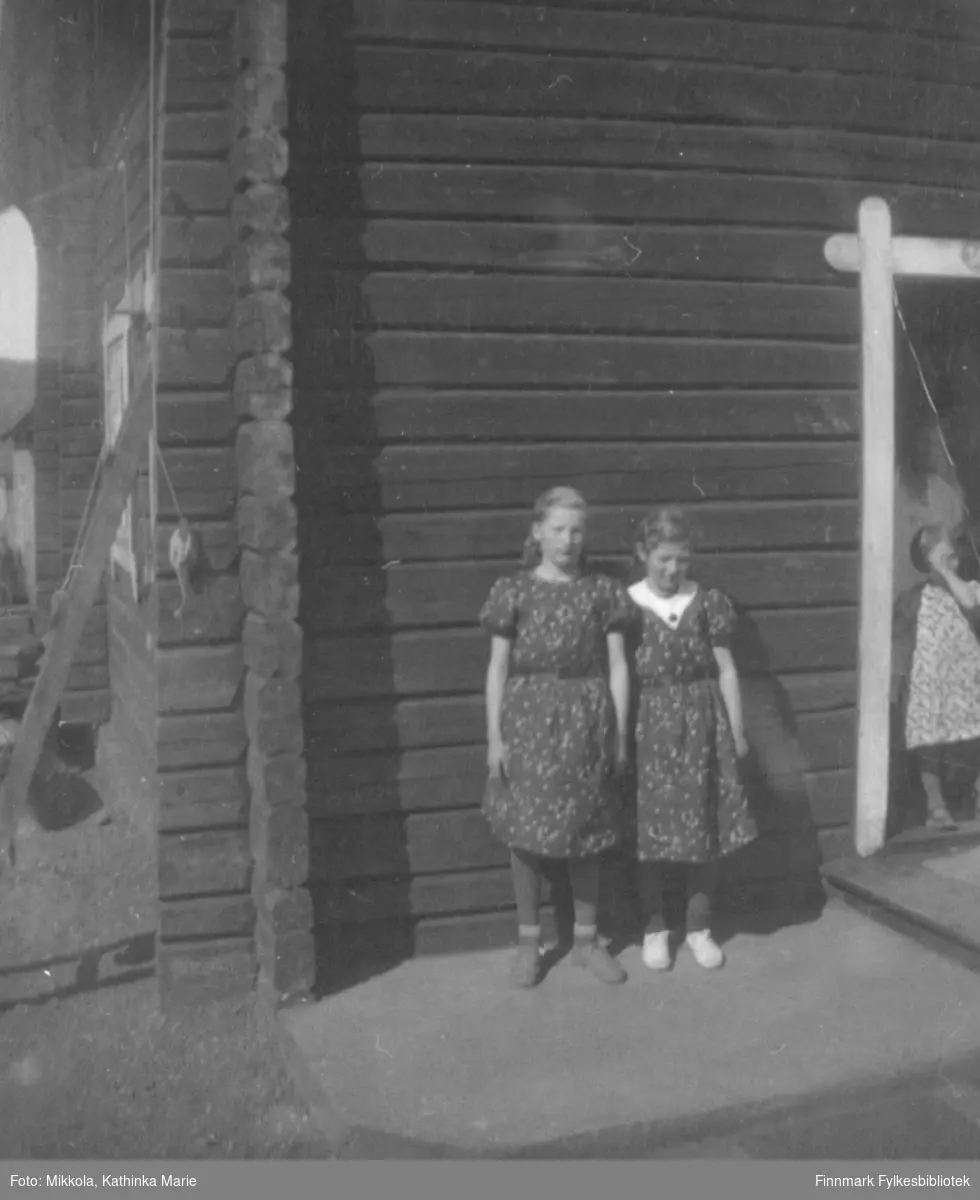 Gudrun (til venstre) og Kari Mikkola fotografert utenfor et tømmerhus i Kirkenes. Dette er samme bygning som på bildene 05007-314, 05007-446 og 05007-478. Ei ukjent, yngre jente står i døråpningen