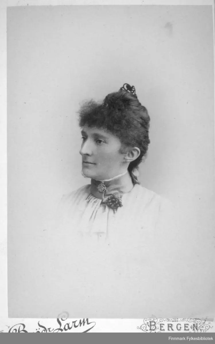 Portrett av en dame i hvit bluse. Hun har håret knytt sammen med et hårbånd på toppen av hodet. Hun har et pyntebånd rundt halsen. Det har en brosje og noe som ser ut som en liten blomsterpynt. Portrettet er tatt av Brødrene Larm i Bergen i februar 1889