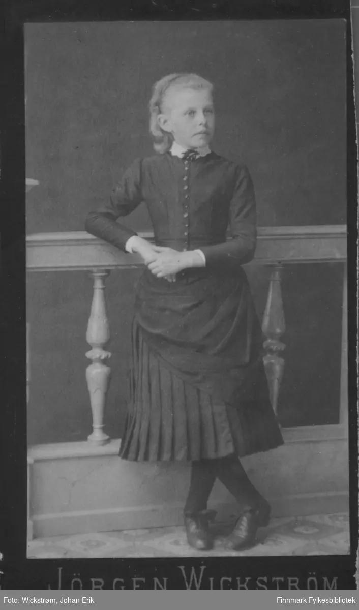 Portrett av en ung jente. Hun har en mørk kjole, mørk strømpebukse og mørke sko på seg. Hun hviler høyrearmen på et rekkverk som har stolper med dreiemønster.