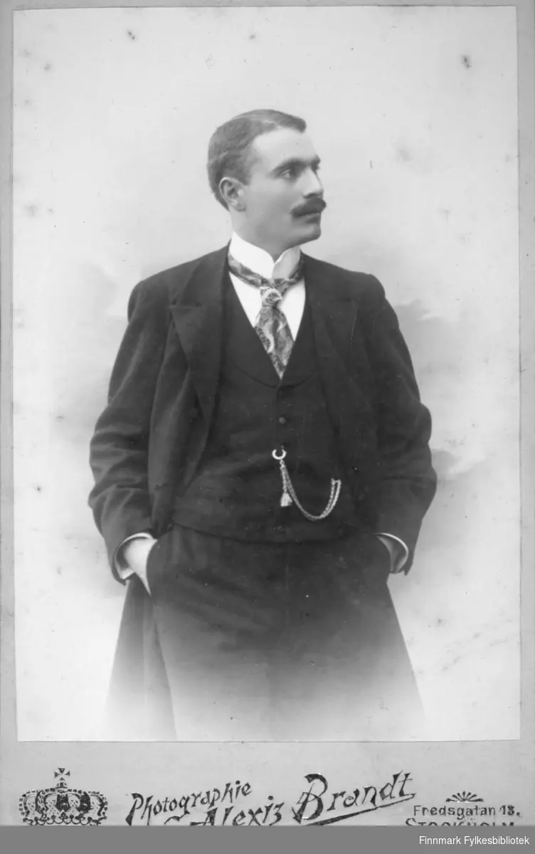 Portrett av en mann iført en mørk dress og frakk. Han har også hvit skjorte og et mønstret slips. Et lommeur-kjede henger fra vestlomma til et knappehull.