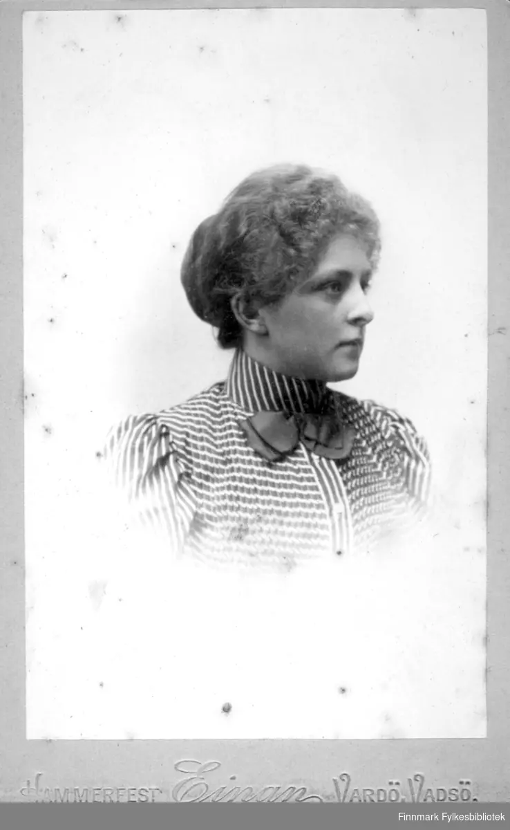 Portrett av en kvinne iført en bluse med striper og mønster. Blusen har en høy krage og en mørk, ensfarget sløyfe er festet i kragen foran.