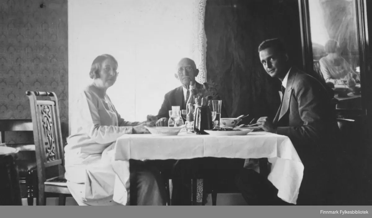 Tre personer, en kvinne og to menn, sitter ved et bord på hotellet i Bossekop. Damen til venstre på bildet er Gerda Buck. Hun har lyst helskjørt og sitter på en stol med utskjæringer på ryggen. Mennene har mørke dresser, skjorte og slips. I midten sitter Hans Ulich som er på norgesbesøk fra USA. En hvit duk ligger på bordet og oppå står kopper og glass, fat, et liten brødkurv, en flaske og en sort kanne. Til høyre henger et stort speil på veggen og midt på bildet er et vindu med lyse, hvite gardiner. Veggene har tapet med vertikalt mønster.