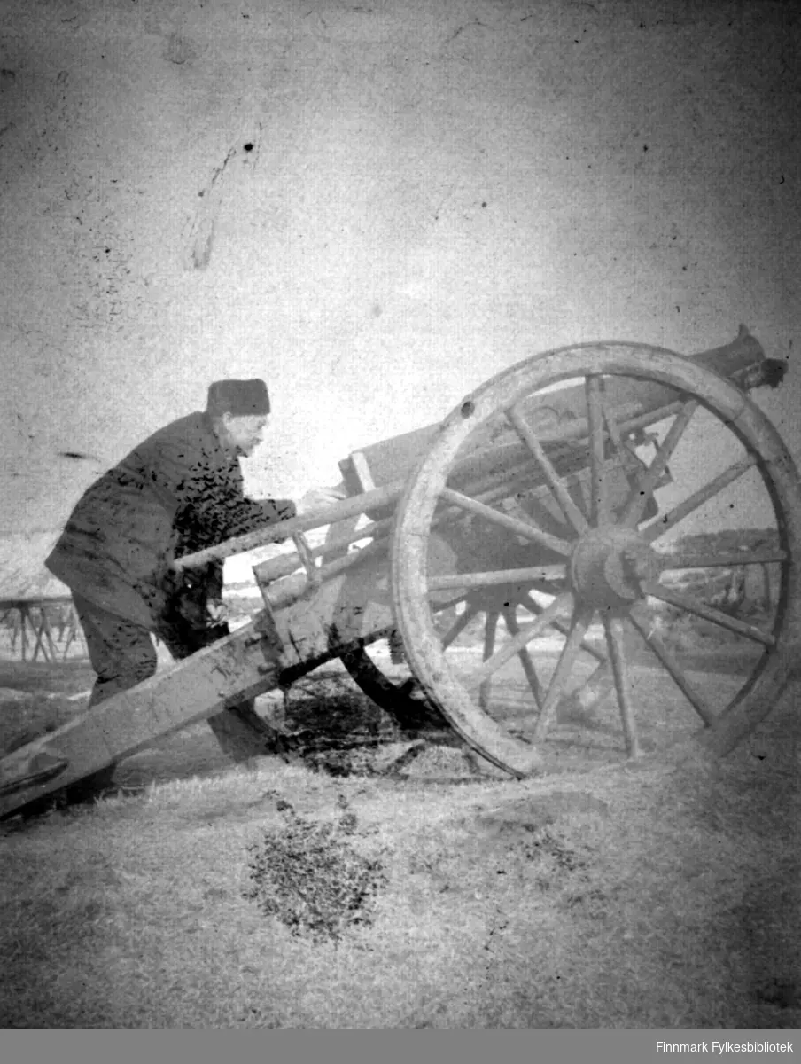 En mann i mørke klær og lue på hodet står bøyd bak en kanon, muligens en Krupp feltkanon. Kanonen har trestativ med lang 'hale' og store trehjul. Det er gress og lyng på bakken og noen snøflekker kan ses i terrenget i bakgrunnen.