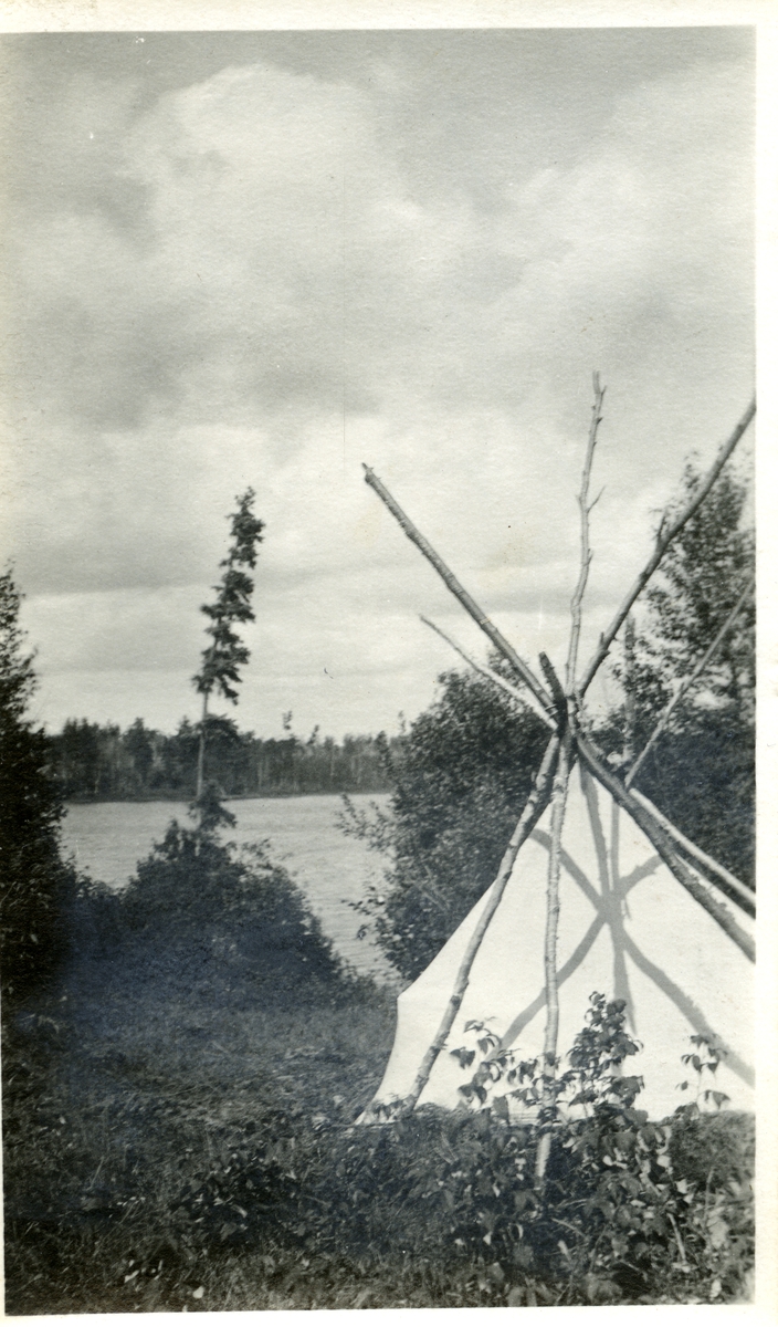 BIldet viser et telt og en uferdig tipi ved bredden av et vann eller en elv.