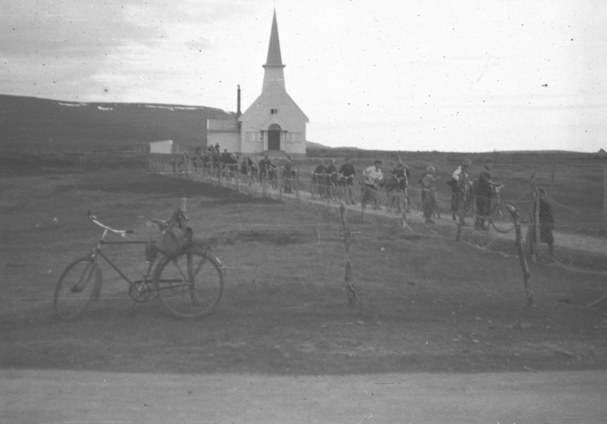 En del personer kommer ned kirkebakken, noen med sykler. Stedet er ukjent.