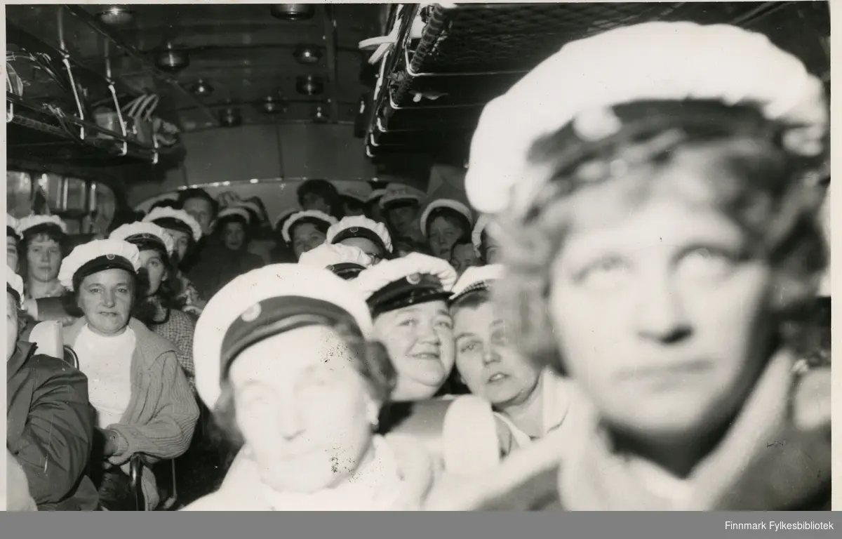 Vadsø damekor i bussen på tur til Berlevåg (muligens rundt 17. mai) 1962. Kvinnene har på seg en hvit hatt, trolig kor uniform. I bussens håndbagasje hylle kan man se vesker og kofferter av forskjellige slag. Damene har på seg jakker og gensere.