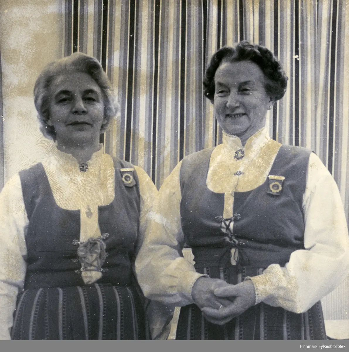 Borghild Dahl Løkting og Klara Pettersen (f. Pedersen) poserer sammen. De to var de som var med og startet Vadsø damekor 16.12.1946. Bildet er trolig tatt på 70/80-tallet. Kvinnene har på seg bunad, smykker og brosjer. Bak dem kan man se en gardin.