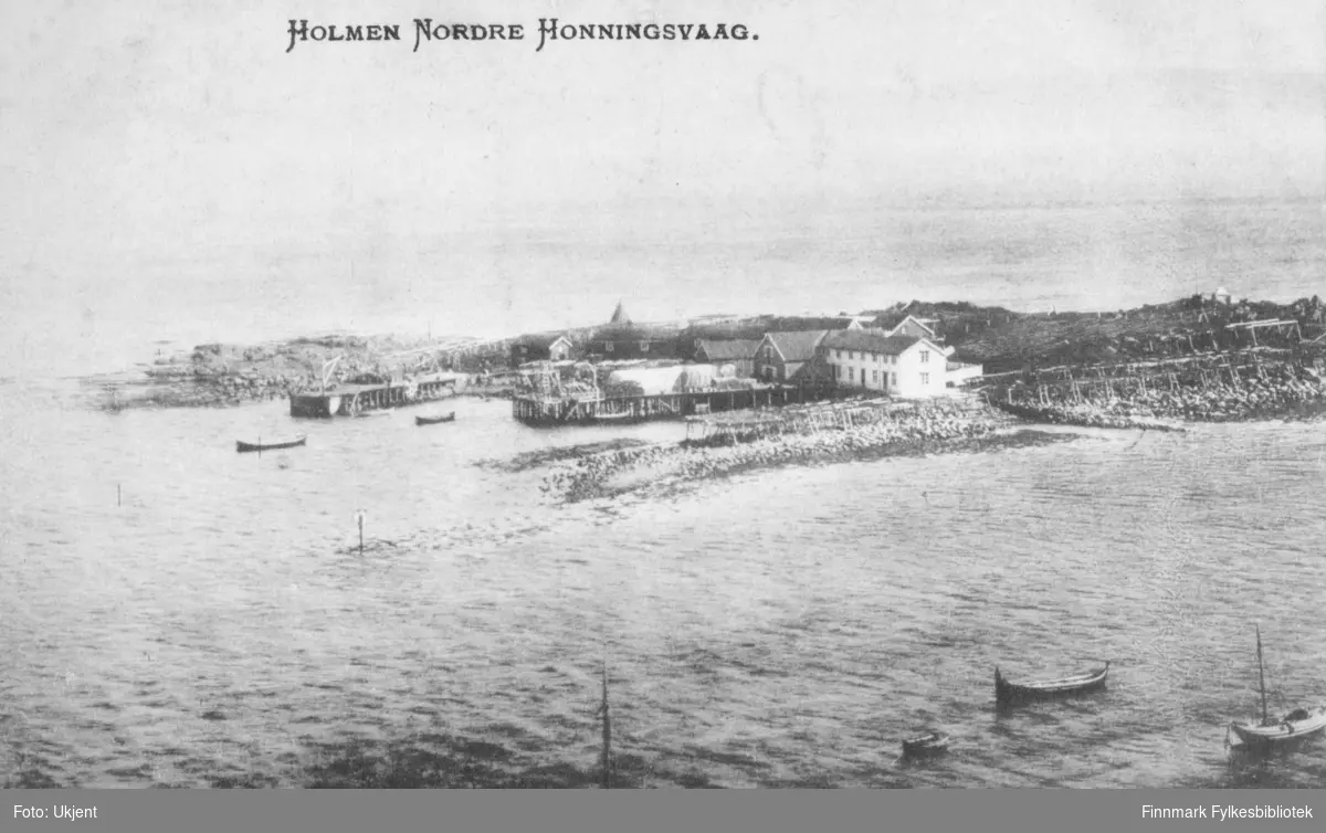 'Holmen Nordre Honningsvaag', står det trykket på dette postkortet. På bilde kan man se en havn med kaier og hus. Enkelte båter ligger ute på havet. I området ligger det flere hjeller.