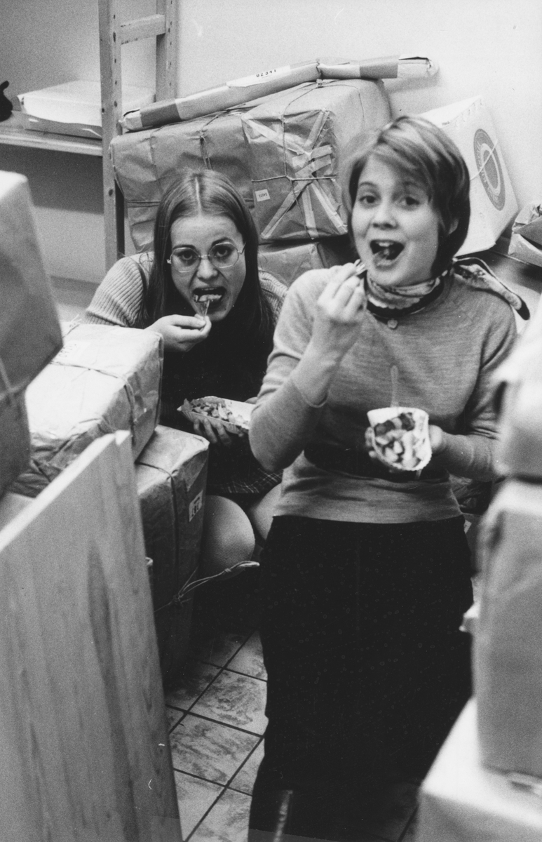 Julruschen på postkontoret Jakobsberg, 1969. Marie-Louise Ericsson och Christina Magnusson, två feriearbetande skolungdomar, tar en paus och äter korv och pommes frites bland alla paketen.