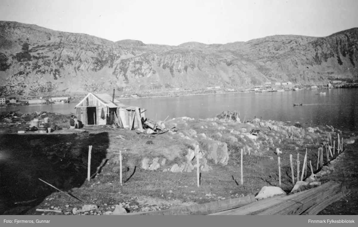 En liten bolig er satt opp av materialer funnet rundt på området. Noen stabler med material ligger i forkant på bildet og boliger står langs strandkanten på andre siden av fjorden. Stedet er ukjent, men bildet er tatt sommeren 1946.