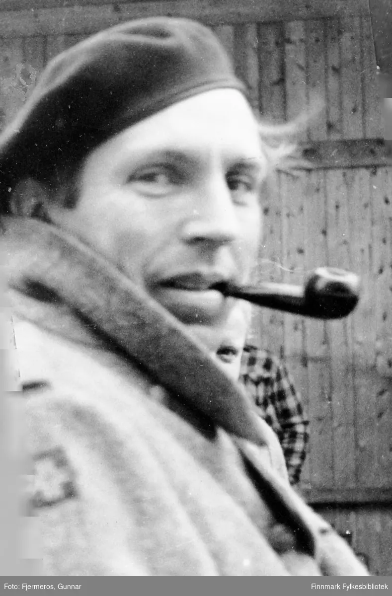 Portrett av en ukjent mann med en tobakkspipe i munnen. To personer til kan såvidt skimtes bak han. Stedet er ukjent, men kan være fra Honningsvåg i 1946.
