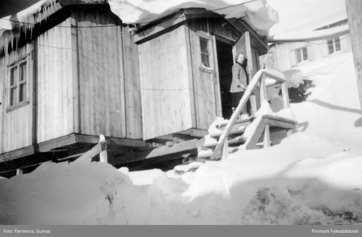 En dame står på trappa til et gjenreisningshus med mye snø på taket og i terrenget. Persoenen er ukjent.