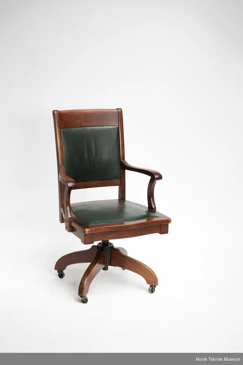 Skrivebordsstol i tre og metall (deler av understellet) med sving, armlener og fire hjul. Sete og rygg i grønt lær.