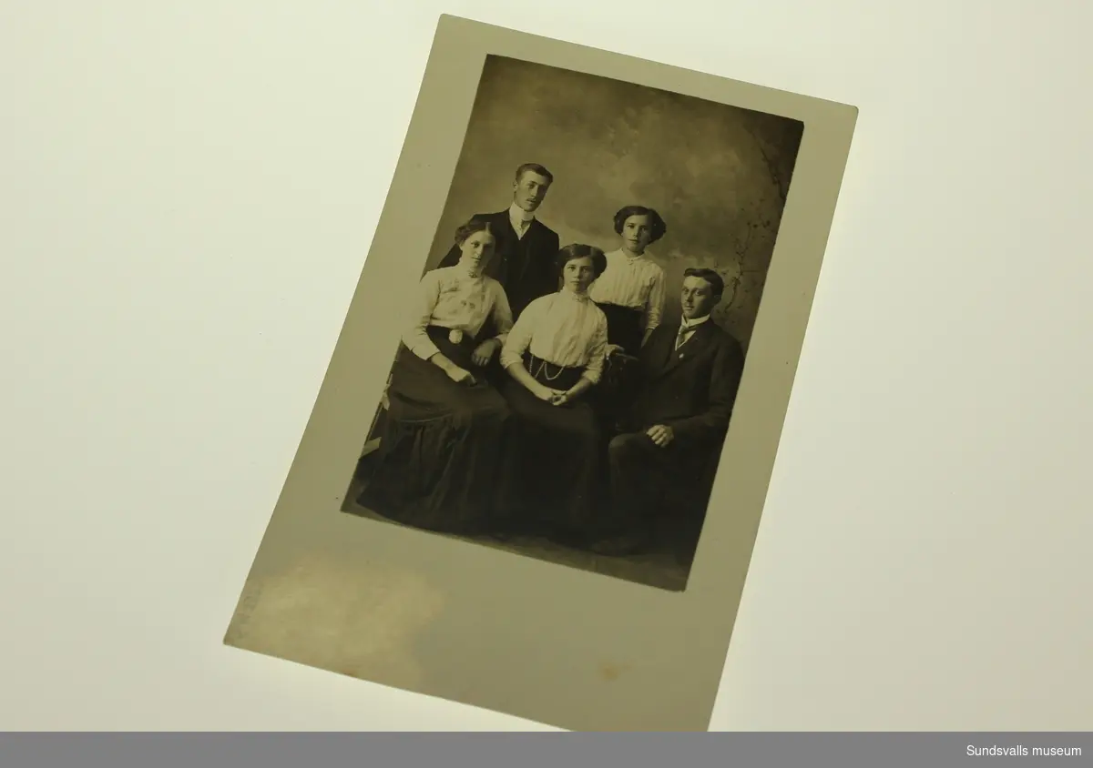 Ett ateljefoto innehållande två kostymklädda män och tre kvinnor samtliga klädda i högt knäppta blusar och långa kjolar.
