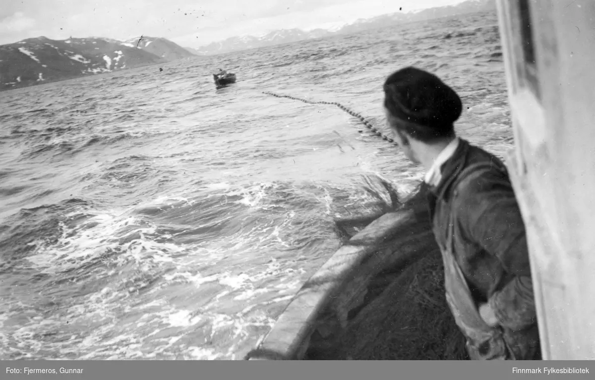En mann står på hekken til en fiskeskøyte som har nota ute, og en lettbåt ligger på vannet i bakgrunnen. Bildet er tatt øst for Nordkapp våren 1948. Personer, sted og båt er ukjent.
