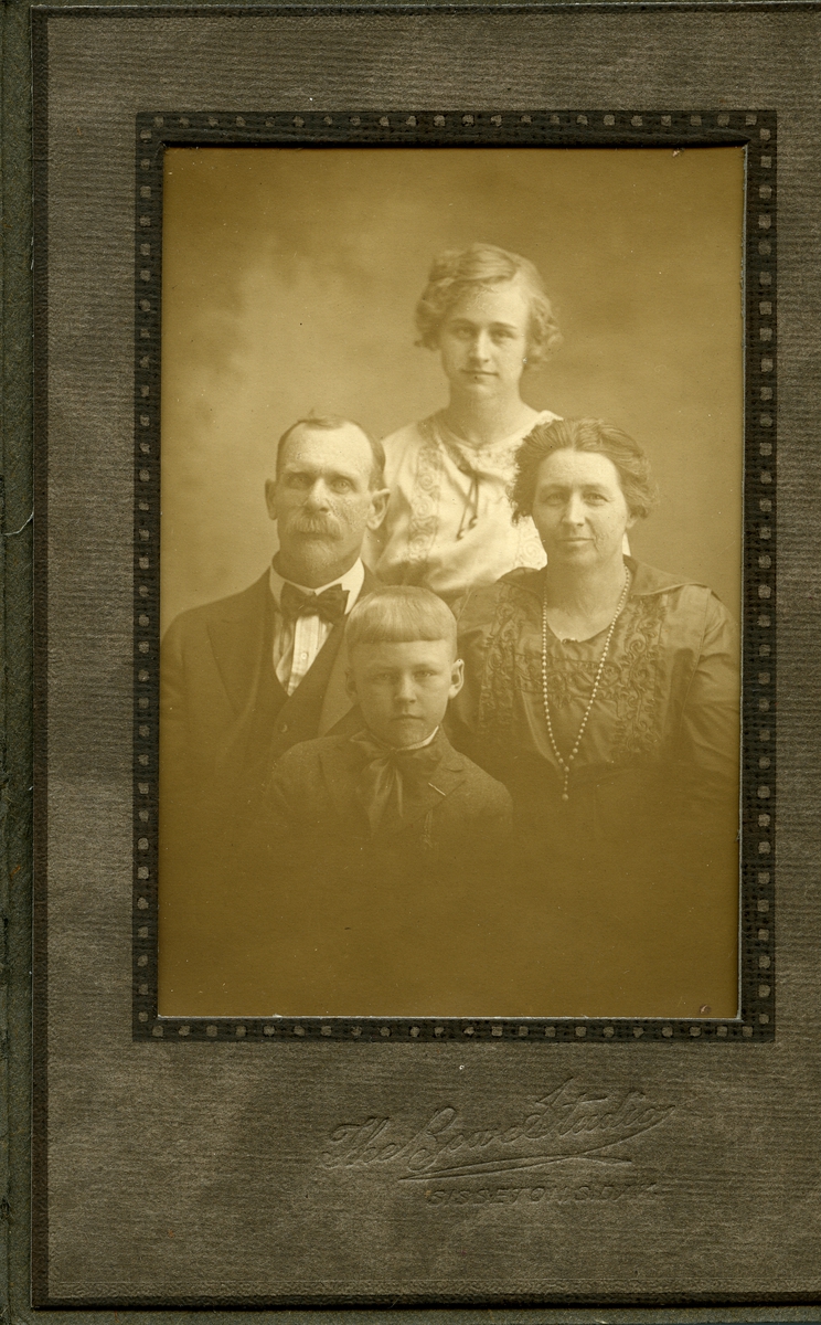 Portrett av en familie foran lerret. Familien består av to barn og mor og far. De er alle ikledd finklær.