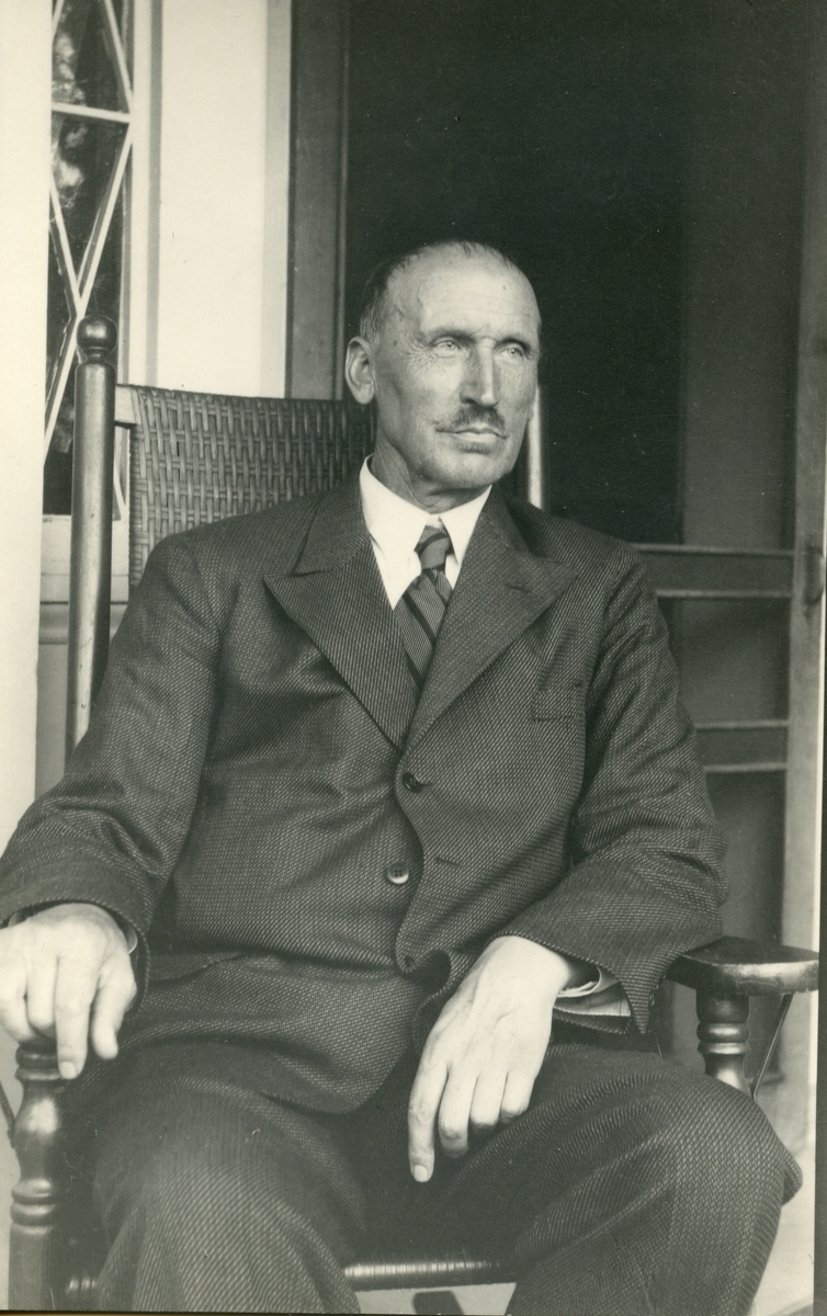 Portrett av en mann iført dress og slips. Mannen sitter i en stol foran en dør med vindu.