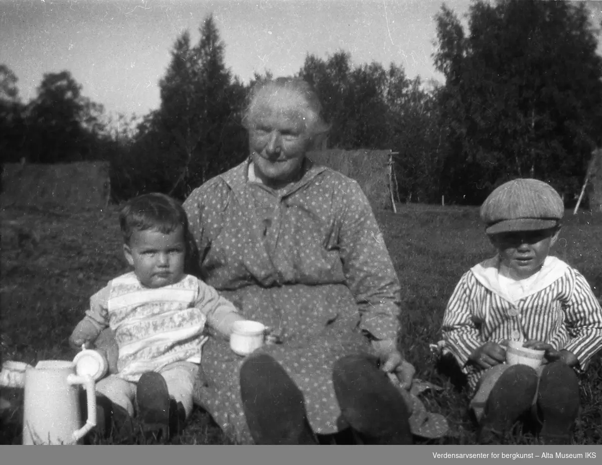 En eldre dame med to små barn på hver side. De sitter på en gressplen.