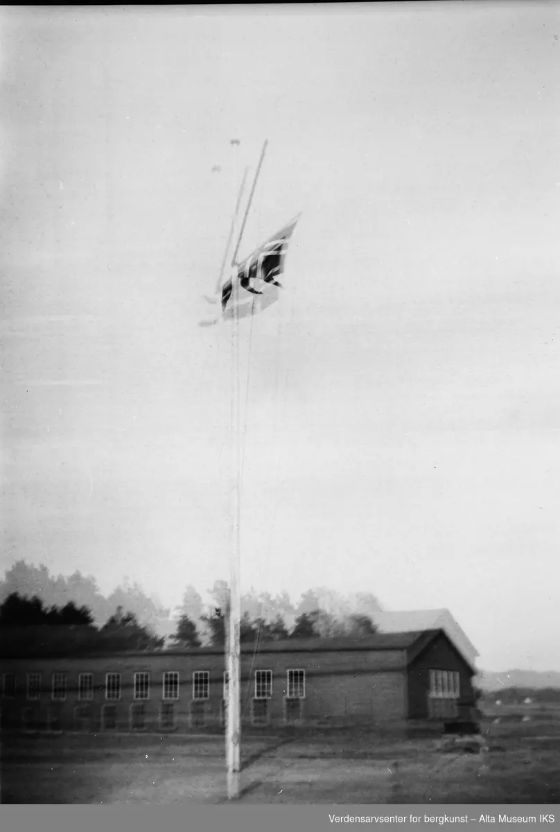 Det norske flagget står heist i flaggstangen foran et langt bygg. Mye bevegelse i bildet.