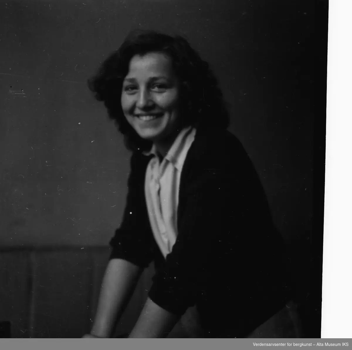 Portrettbilde av en ung kvinne som smiler til kameraet. Hun lener seg mot noe.