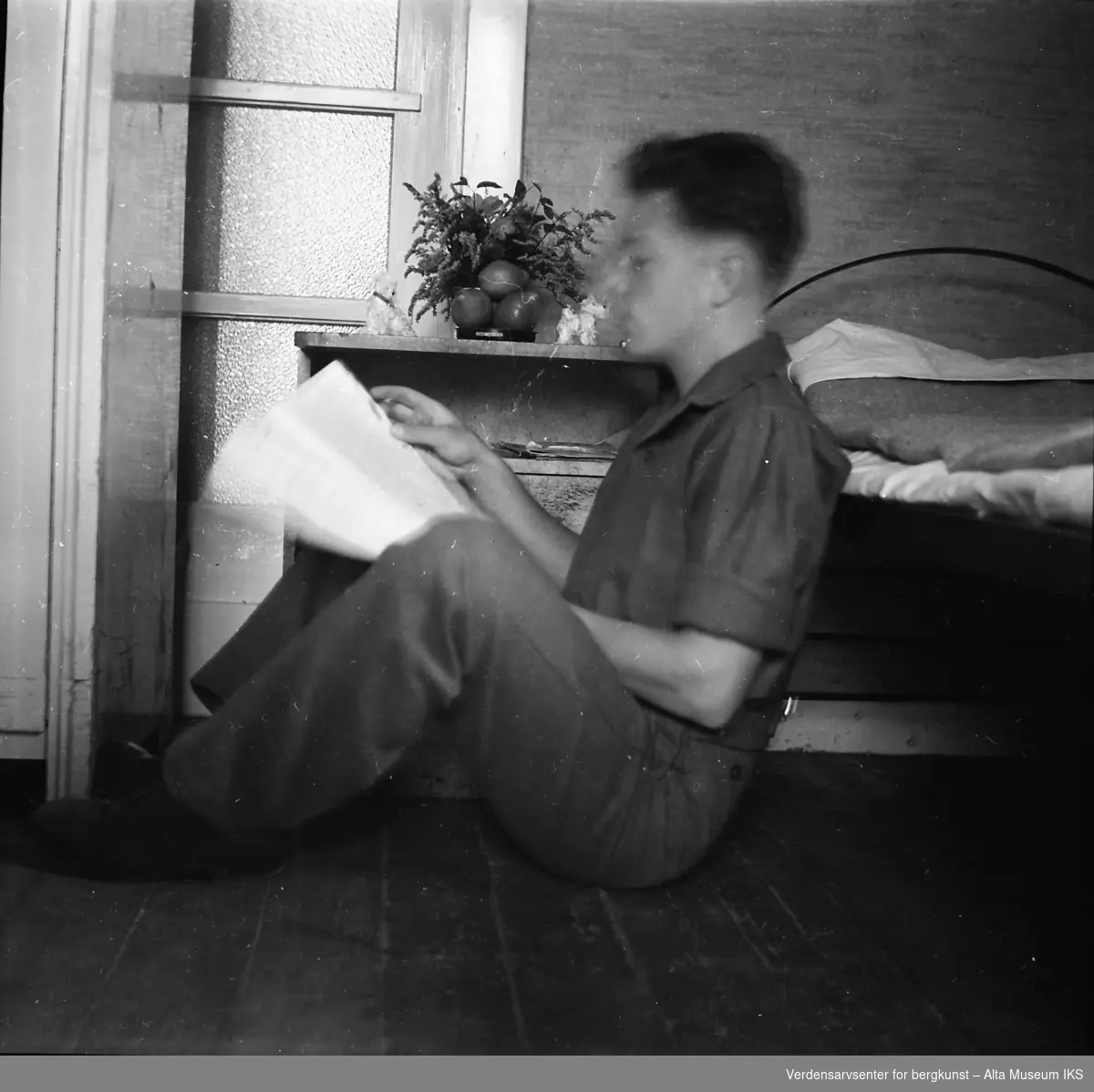 En ung mann sitter på gulvet og lener seg mot en seng. Han leser en bok.