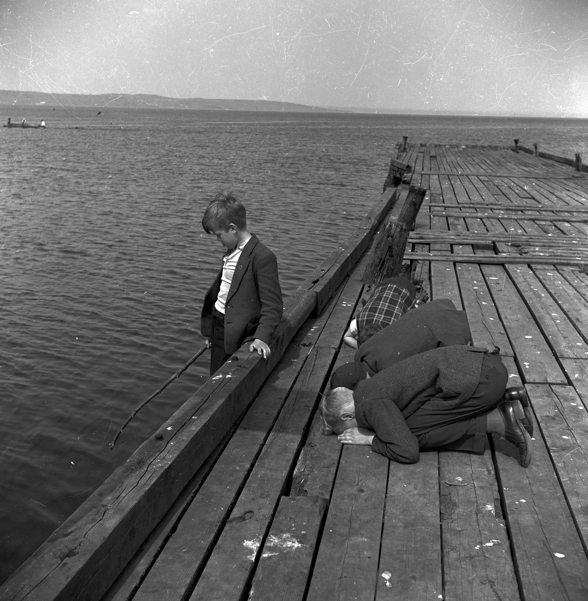 På en brygga ligger tre pojkar på knä och kikar ner mellan springorna. Vid sidan om står en pojke och fiskar med metreven knuten om en pinne.