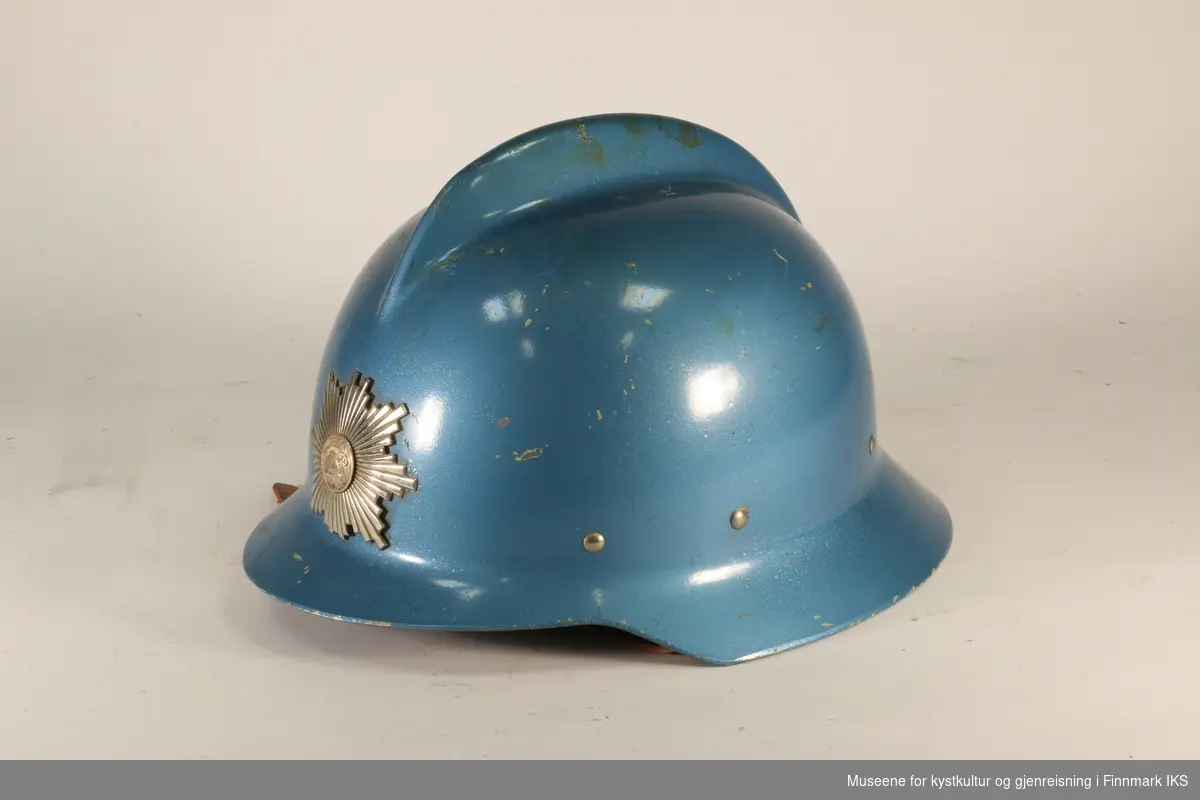 Blå brannhjelm av metall med hanekam på toppen. Foran er det brannvesenets emblem. Innerfor av hjelmen er av skinn. Foret kan reguleres med snor.