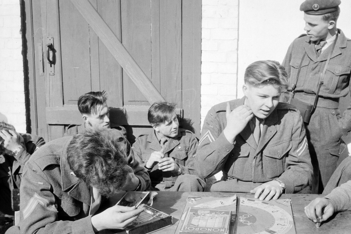 Soldater slapper av i solvegg. I forgrunnen et "Monopol"-spill på et bord.